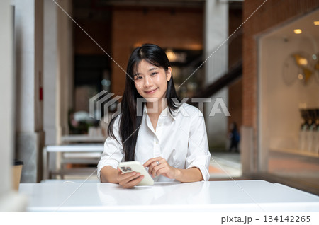 Asian young woman holding or using smartphone while sitting at table aside coffee cafe in a building 134142265