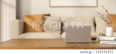 Laptop and a glass of coffee on wooden table with books and vase across sofa in blurred living room. Laptop and a glass of coffee on wooden table with books and vase across sofa in blurred living room. 134142351