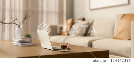Laptop and a glass of coffee on wooden table with books and vase aside sofa in a blurred living room 134142357