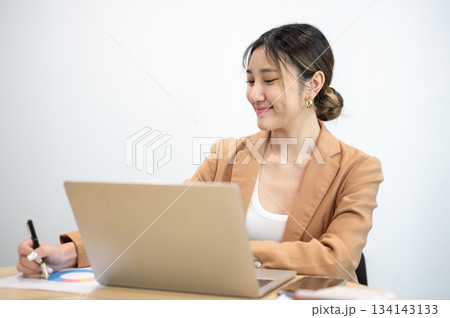 Asian businesswoman office worker holding a pen under chain looking at laptop on table in white room Asian businesswoman office worker holding a pen under chain looking at laptop on table in white room 134143133
