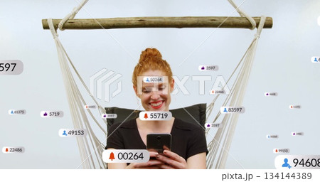 Smiling red-haired woman holding phone in rope wooden hammock chair at studio with notifications Smiling red-haired woman holding phone in rope wooden hammock chair at studio with notifications 134144389