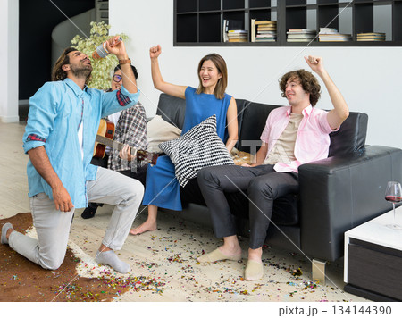 Group of young multiracial people having fun at house party. Young friends playing guitar and singing while sitting on sofa in living room. Group of young multiracial people having fun at house party. Young friends playing guitar and singing while sitting on sofa in living room. 134144390
