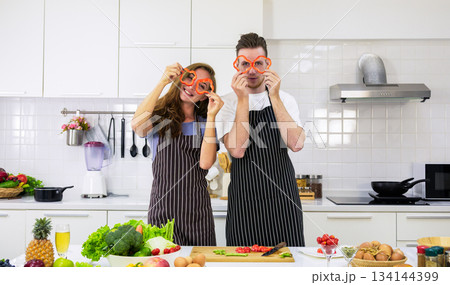 Playful couple wearing black striped aprons holding red bell pepper rings as glasses over eyes in modern kitchen. Fresh vegetables and fruit arranged on counter at home Playful couple wearing black striped aprons holding red bell pepper rings as glasses over eyes in modern kitchen. Fresh vegetables and fruit arranged on counter at home 134144399