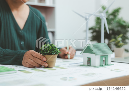 Eco-friendly housing model with solar panel and business charts on desk, symbolizing green building, renewable energy, sustainable architecture and environmental innovation concept. Eco-friendly housing model with solar panel and business charts on desk, symbolizing green building, renewable energy, sustainable architecture and environmental innovation concept. 134146622