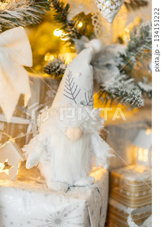 White troll figure under snow covered Christmas tree with white decoration. White, silver and gold gift boxes. New Year, Christmas. Warm light 134151222