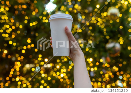 Woman hand holding disposable takeaway coffee cup with Christmas tree background outdoors. Mockup, space for text. Blurred background with Christmas lights. Recyclable eco packaging. 134151230