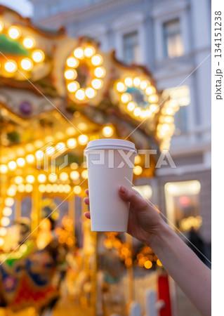 Woman hand holding disposable takeaway coffee cup with Christmas carousel on Christmas fair in the background. Mockup, space for text. Blurred background. Recyclable packaging 134151238