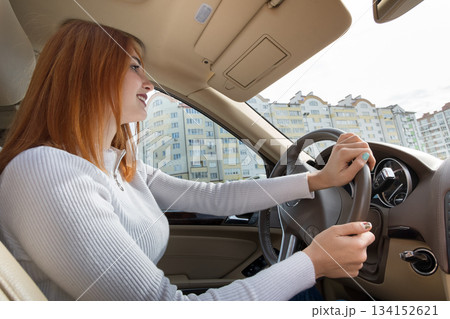 Young redhead woman driver behind a wheel driving a car smiling happily. 134152621