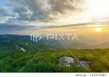 Wide summer mountain view at sunrise. Glowing orange sun raising in blue cloudy sky over green grassy hill with big rock and distant mountain range covered with morning mist. Beauty of nature concept. Wide summer mountain view at sunrise. Glowing orange sun raising in blue cloudy sky over green grassy hill with big rock and distant mountain range covered with morning mist. Beauty of nature concept. 134152663