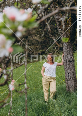 Smiling middle-aged woman standing near blooming tree in spring garden. Concept of female health, hormonal balance, wellness, and natural beauty in midlife Smiling middle-aged woman standing near blooming tree in spring garden. Concept of female health, hormonal balance, wellness, and natural beauty in midlife 134154451