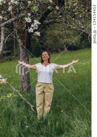Smiling middle-aged woman standing near blooming tree in spring garden. Concept of female health, hormonal balance, wellness, and natural beauty in midlife 134154453