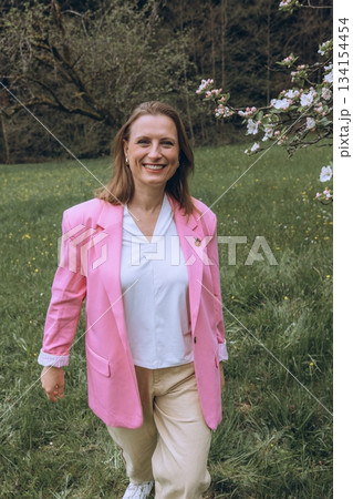 Smiling middle-aged woman standing near blooming tree in spring garden. Concept of female health, hormonal balance, wellness, and natural beauty in midlife 134154454