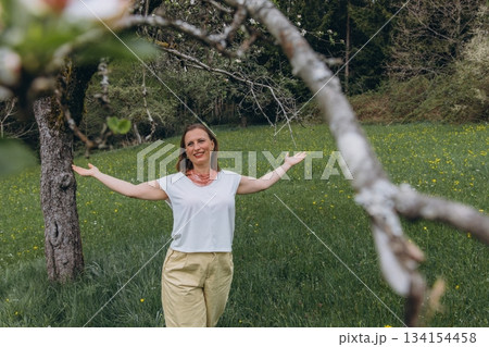 Smiling middle-aged woman standing near blooming tree in spring garden. Concept of female health, hormonal balance, wellness, and natural beauty in midlife Smiling middle-aged woman standing near blooming tree in spring garden. Concept of female health, hormonal balance, wellness, and natural beauty in midlife 134154458