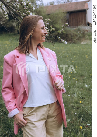 Smiling middle-aged woman standing near blooming tree in spring garden. Concept of female health, hormonal balance, wellness, and natural beauty in midlife Smiling middle-aged woman standing near blooming tree in spring garden. Concept of female health, hormonal balance, wellness, and natural beauty in midlife 134154464