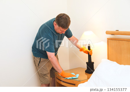 Man in Casual Attire Dusting Under a Desk Lamp, Embracing His Role as a Stay-at-Home Husband in a Tidy and Inviting Living Space Man in Casual Attire Dusting Under a Desk Lamp, Embracing His Role as a Stay-at-Home Husband in a Tidy and Inviting Living Space 134155377