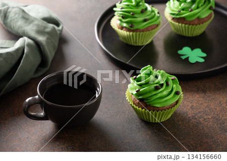 St Patricks day chocolate cupcakes with green cream frosting and coffee cup on dark table. Festive dessert concept for Irish holiday. 134156660