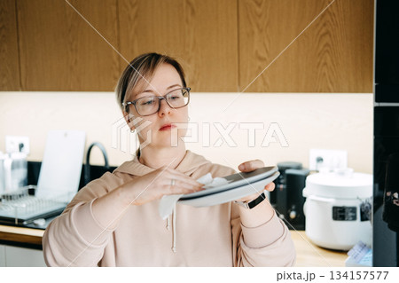 A woman wipes a gray plate with a small white cloth in a modern kitchen. Sustainable kitchen alternatives, reducing paper waste, reusable cloths, eco-friendly cleaning A woman wipes a gray plate with a small white cloth in a modern kitchen. Sustainable kitchen alternatives, reducing paper waste, reusable cloths, eco-friendly cleaning 134157577