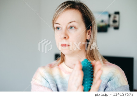 A woman holds a spiky massage ball in both hands and looks off to the side, contemplating health routine. Accessible health care, budget wellness, DIY health solutions, proactive health maintenance. A woman holds a spiky massage ball in both hands and looks off to the side, contemplating health routine. Accessible health care, budget wellness, DIY health solutions, proactive health maintenance. 134157578