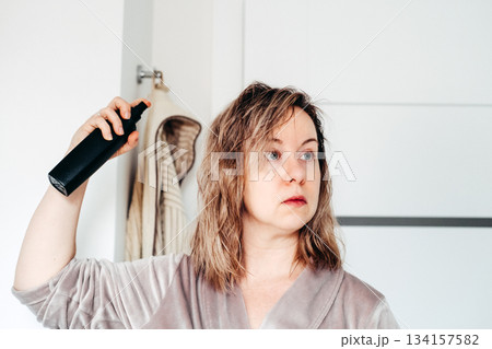 A woman in a robe sprays a product onto her damp shoulder length hair, looking into the distance with a serious expression. Hair loss concern, hair maintenance, clean haircare, self appraisal. 134157582
