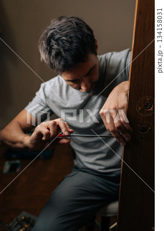 Vertical shot of professional technician male fixing door lock with screwdriver, performing installation and repair work on mechanism to ensure security and functionality within house. 134158031