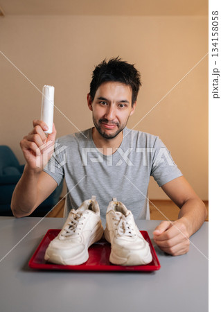 Vertical portrait of smiling man holding white sneaker and shoe cleaner bottle, preparing to clean footwear at home, demonstrating process of refreshing shoes and maintaining appearance. Vertical portrait of smiling man holding white sneaker and shoe cleaner bottle, preparing to clean footwear at home, demonstrating process of refreshing shoes and maintaining appearance. 134158058