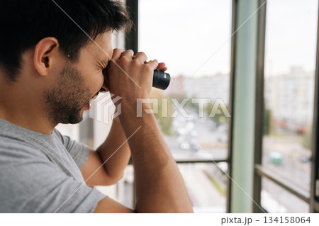 Curious young adult man holding monocular, looking closely out of window at urban street, engaging in observation, surveillance and spying while monitoring surroundings from apartment or home. 134158064