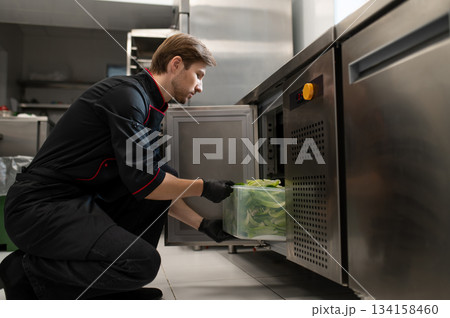 Male cook places large container of lettuce placed into refrigerator 134158460