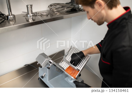 Man in uniform slicing meat with professional slicer 134158461