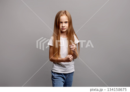 Aggressive young child girl clenching fists in emotional defensive pose, expressing frustration and defiance in studio portrait on gray background, showing intense feelings and reaction 134158675