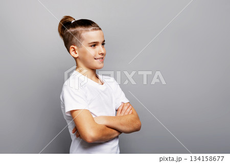 Stylish young boy posing in studio with trendy shaved sides and sleek ponytail hairstyle, smiling with arms crossed, modern fashion portrait on clean gray background 134158677