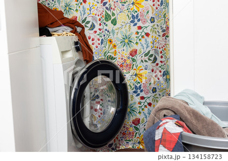 A washing machine sits in a laundry space surrounded by a bright floral wall design. Clothes are piled nearby, creating a busy scene of household chores A washing machine sits in a laundry space surrounded by a bright floral wall design. Clothes are piled nearby, creating a busy scene of household chores 134158723