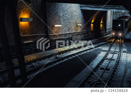 Dimly lit tunnel reveals steep Heidelberg funicular tracks curving toward arrive carriage with bright headlights. Cool tones warm lamps create dramatic contrast industrial underground transport scene Dimly lit tunnel reveals steep Heidelberg funicular tracks curving toward arrive carriage with bright headlights. Cool tones warm lamps create dramatic contrast industrial underground transport scene 134158828