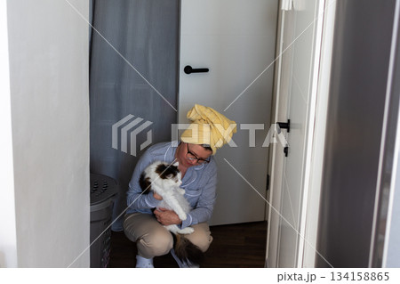 A person is sitting on the floor of a hallway holding a white and brown cat. They have a towel on their head and are dressed in casual clothes. It is daytime and bright inside 134158865