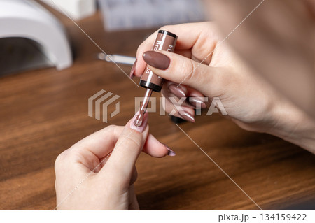 A woman applies brown gel nail polish to her own long nails during a home manicure session, emphasizing personal care and beauty routine A woman applies brown gel nail polish to her own long nails during a home manicure session, emphasizing personal care and beauty routine 134159422