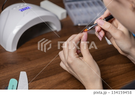 Woman applying polygel onto a nail tip for a DIY manicure by oneself at home, utilizing a UV lamp for a beauty routine 134159458