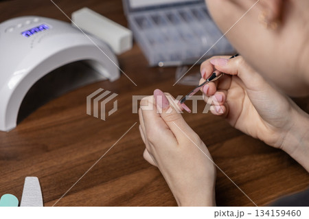 Woman's hands applying gel nail extension by oneself using a tool, performing a DIY home manicure with a UV lamp for personal beauty Woman's hands applying gel nail extension by oneself using a tool, performing a DIY home manicure with a UV lamp for personal beauty 134159460