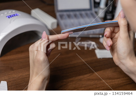 Woman meticulously files her own long acrylic nail extension, with a UV lamp nearby, highlighting personal beauty care and a precise DIY manicure routine Woman meticulously files her own long acrylic nail extension, with a UV lamp nearby, highlighting personal beauty care and a precise DIY manicure routine 134159464