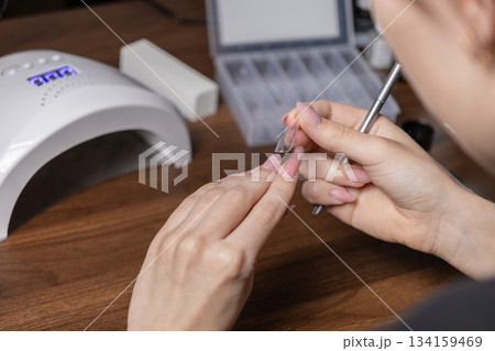 Adult woman performs self-manicure, carefully applying a clear nail extension upper form with pink gel, utilizing a modern UV LED lamp and essential beauty tools 134159469