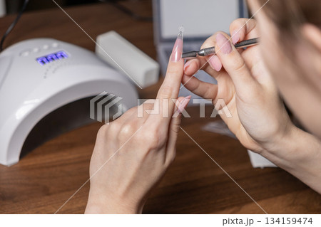 Young woman carefully performing a DIY nail extension manicure on herself, applying an upper form with a tool for perfect, elegant long nails 134159474