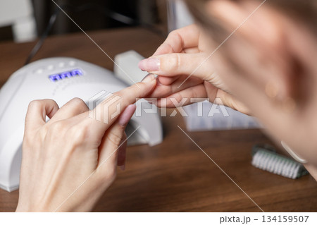 Woman skillfully applies her own gel nail extension using modern upper forms and a professional UV lamp, perfecting a DIY manicure for self-care beauty 134159507