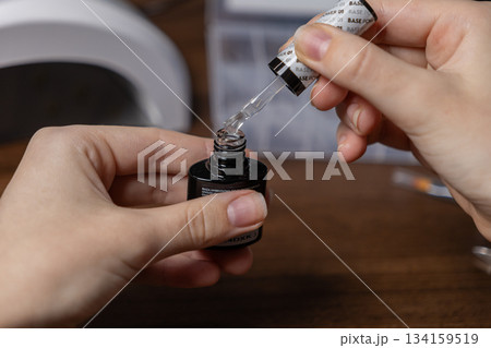 A person's hands carefully dipping a nail polish brush into a transparent gel lacquer bottle, signifying a self-care beauty routine or home manicure preparation A person's hands carefully dipping a nail polish brush into a transparent gel lacquer bottle, signifying a self-care beauty routine or home manicure preparation 134159519