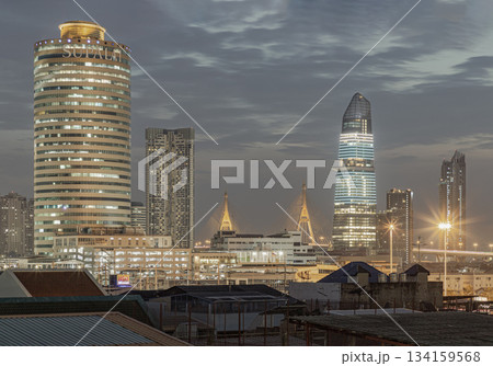 Under a dramatic, cloudy sky, the illuminated KingBridge tower and other modern skyscrapers stand as a stunning and iconic landmark of Bangkok's business district, alongside the majestic Bhumibol 134159568