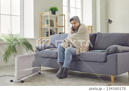 Man sitting at home wrapped in blanket talking by mobile phone warming up with electric heater. Man sitting at home wrapped in blanket talking by mobile phone warming up with electric heater. 134160178