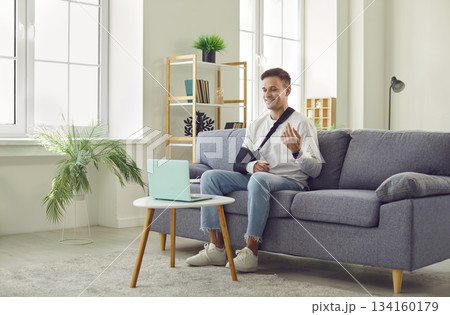 Young man with arm in cast sitting on couch at home and and having online video call on a laptop. Young man with arm in cast sitting on couch at home and and having online video call on a laptop. 134160179