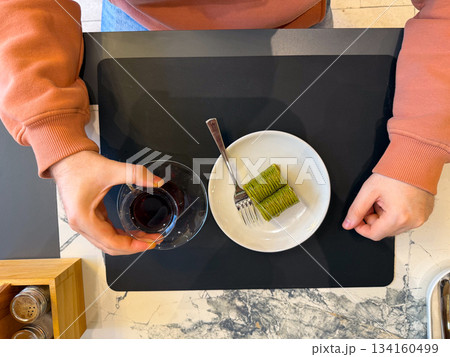 Hands with tea and pistachio baklava from top view. Cozy cafe break, sweet dessert moment, warmth, calm dining atmosphere and gentle pause. 134160499