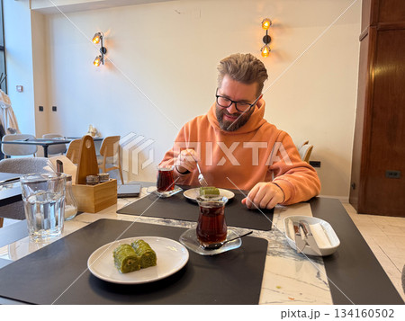 Smiling man with baklava and tea on the table. Dining joy, relaxed break, warm cafe atmosphere, sweetness, communication and comfort. 134160502