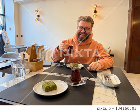 Man enjoying Turkish pistachio baklava at cafe table. Warm dining scene, dessert tasting moment, hospitality, comfort, tea break and joyful atmosphere. Man enjoying Turkish pistachio baklava at cafe table. Warm dining scene, dessert tasting moment, hospitality, comfort, tea break and joyful atmosphere. 134160503