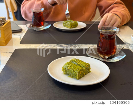 Two plates of Turkish pistachio baklava served with tea. Warm cafe moment, social dining, shared dessert experience and cozy break together. Two plates of Turkish pistachio baklava served with tea. Warm cafe moment, social dining, shared dessert experience and cozy break together. 134160506