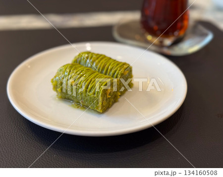 Pistachio baklava dessert on white plate close-up. Cafe sweetness, gourmet treat, green pastry indulgence with tea and cozy dining moment. Pistachio baklava dessert on white plate close-up. Cafe sweetness, gourmet treat, green pastry indulgence with tea and cozy dining moment. 134160508