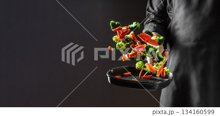 Chef tossing fresh broccoli and carrot slices into a hot grill pan in mid-air action on dark background with copy space for culinary advertising Chef tossing fresh broccoli and carrot slices into a hot grill pan in mid-air action on dark background with copy space for culinary advertising 134160599